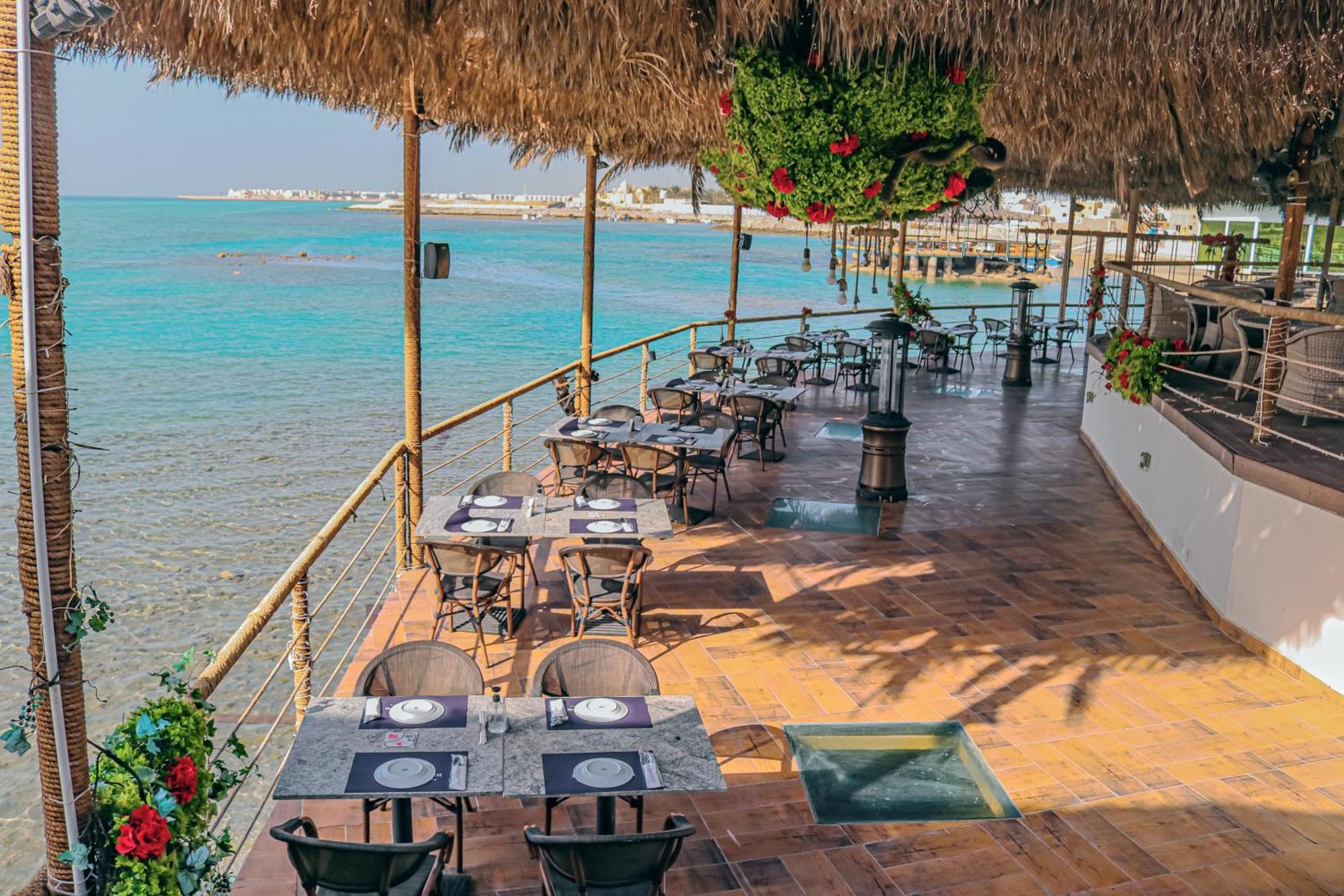 Lagoona Beach Luxury Resort And Spa Manama Luaran gambar The photo shows a seaside restaurant with a spacious outdoor dining area. It features several tables set with plates and utensils, surrounded by lush greenery and decorative plants. The flooring is made of brown tiles, and there are thatched roofs pr