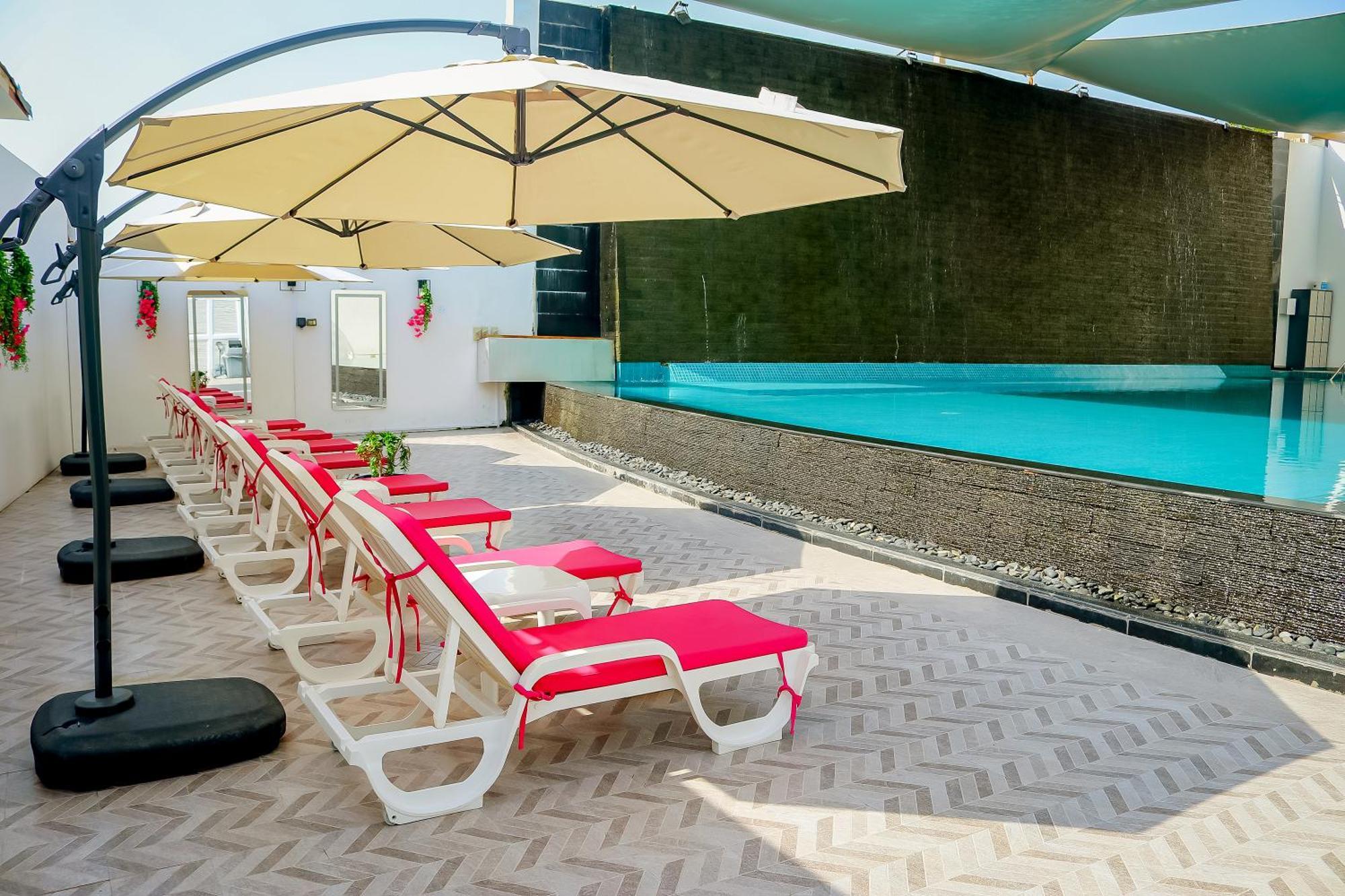 Lagoona Beach Luxury Resort And Spa Manama Luaran gambar The photo shows a sunny poolside area with several lounge chairs arranged in a row. Each chair features red cushions and is positioned under large, beige umbrellas for shade. In the background, there is a beautiful swimming pool with clear blue water