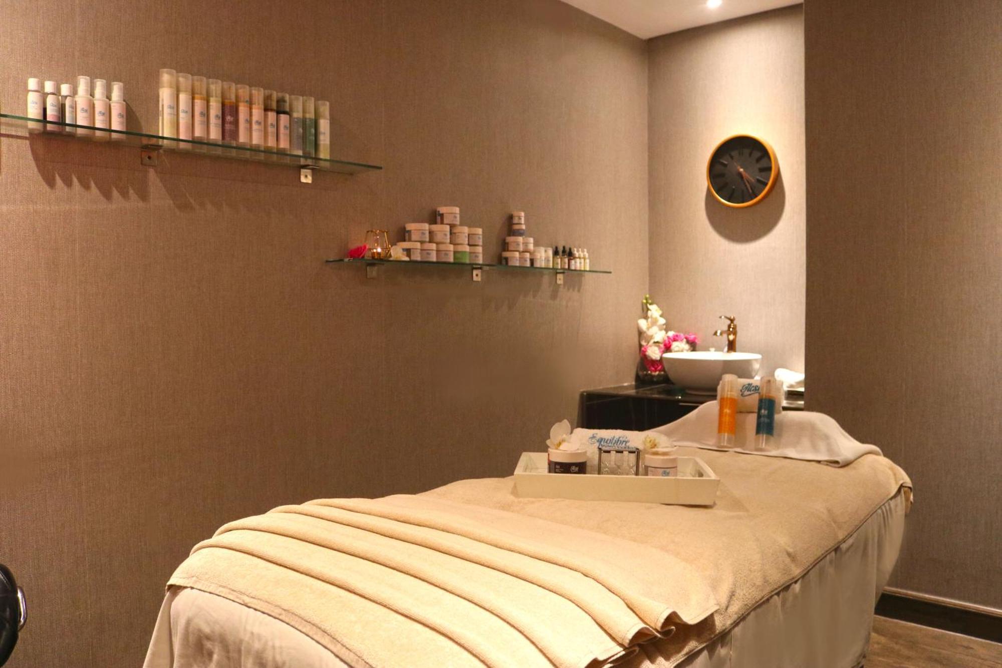 Lagoona Beach Luxury Resort And Spa Manama Luaran gambar The photo shows a spa treatment room. There is a massage table draped with a light-colored blanket, creating a relaxing atmosphere. On the wall, there are shelves displaying various skincare products in bottles and jars. A clock is mounted on the wal
