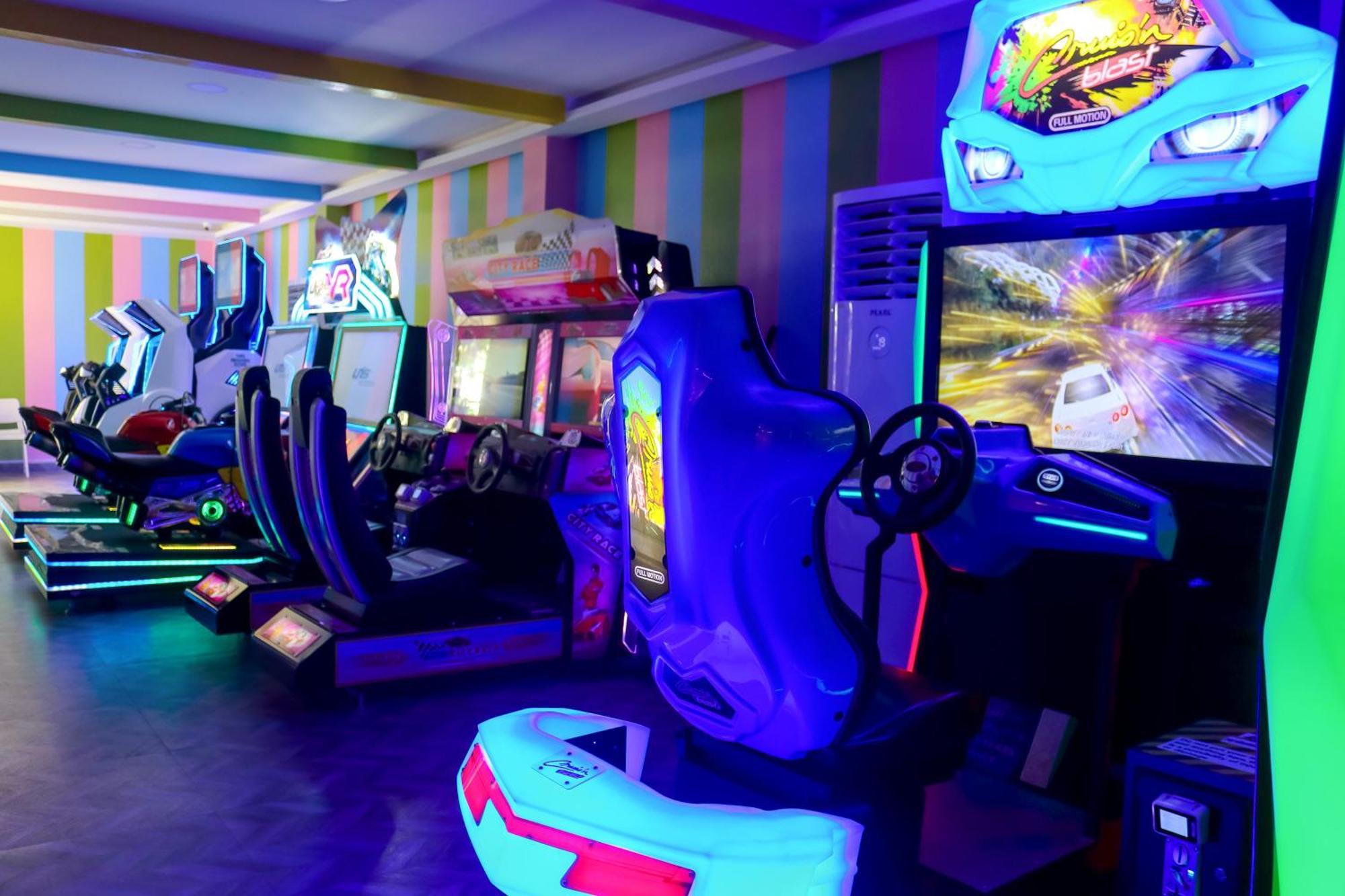 Lagoona Beach Luxury Resort And Spa Manama Luaran gambar The photo shows an arcade area filled with various arcade machines. The setting is illuminated with colorful neon lights, creating a vibrant atmosphere. There are several racing game machines with steering wheels and seats, each accompanied by screen