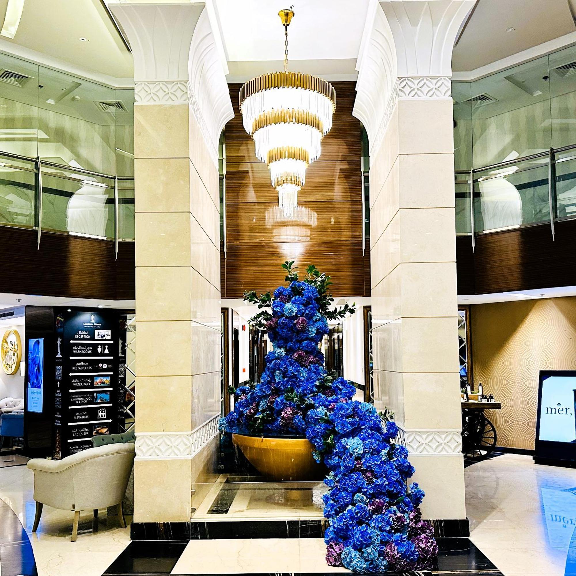 Lagoona Beach Luxury Resort And Spa Manama Luaran gambar The photo depicts an elegant lobby or entrance area of a building. Central to the scene is a large floral arrangement featuring vibrant blue flowers. This arrangement is positioned in a decorative bowl. Above it hangs a stylish chandelier with a laye