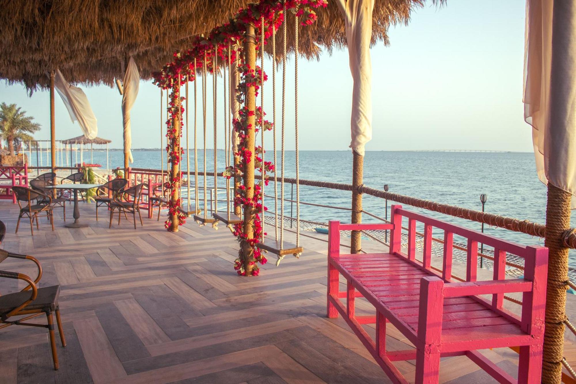 Lagoona Beach Luxury Resort And Spa Manama Luaran gambar The photo depicts a scenic outdoor area by the water, with a wooden deck and a thatched roof. Two swings adorned with flowers are hanging from the ceiling, providing a charming touch to the space. In the foreground, there is a bright pink bench, addi