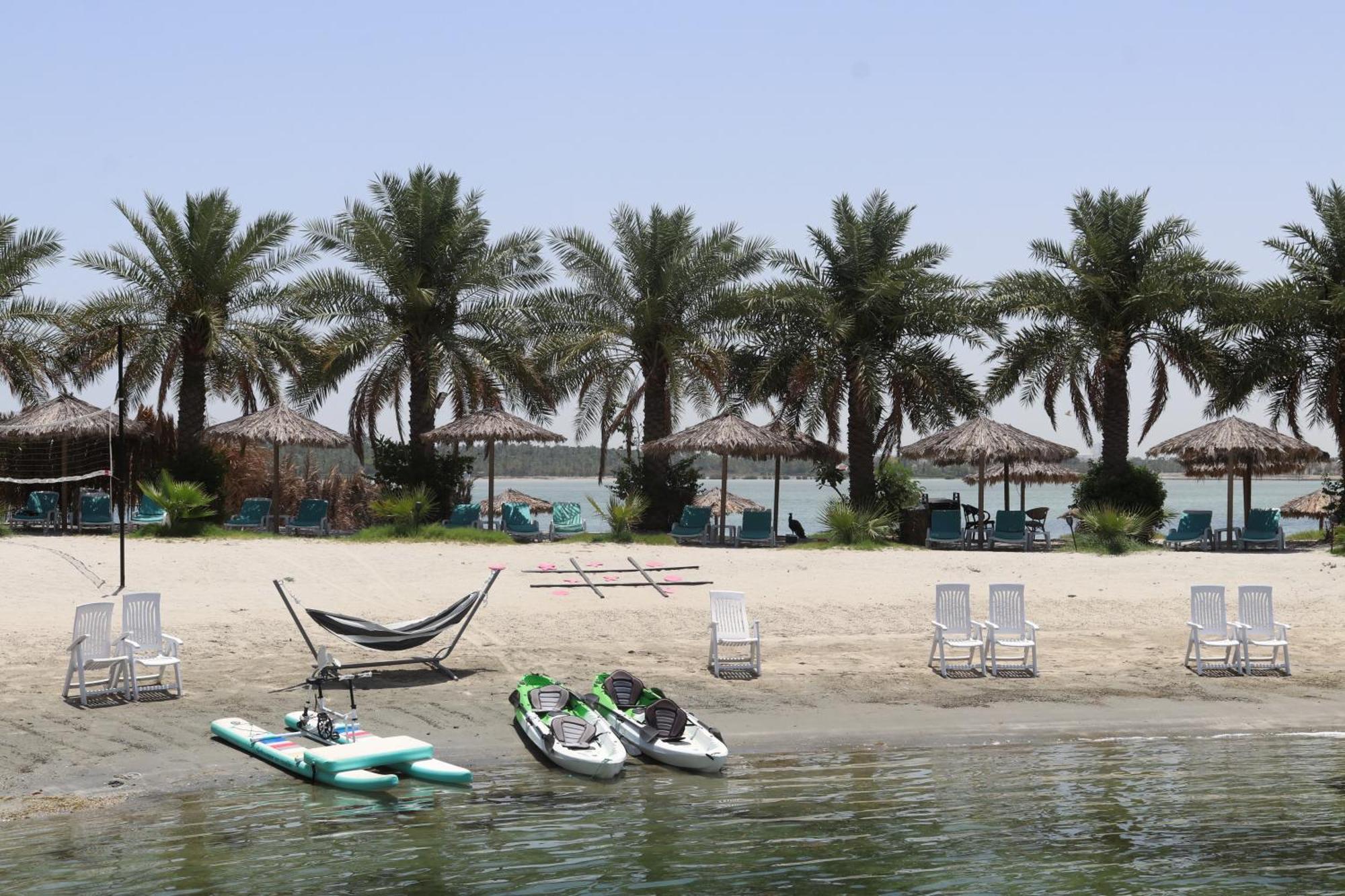 Lagoona Beach Luxury Resort And Spa Manama Luaran gambar The photo depicts a serene beach scene with several palm trees lining the shore. There are thatched umbrellas providing shade, and a few lounge chairs are positioned on the sand. A hammock is set up between two supports, inviting relaxation. In the f