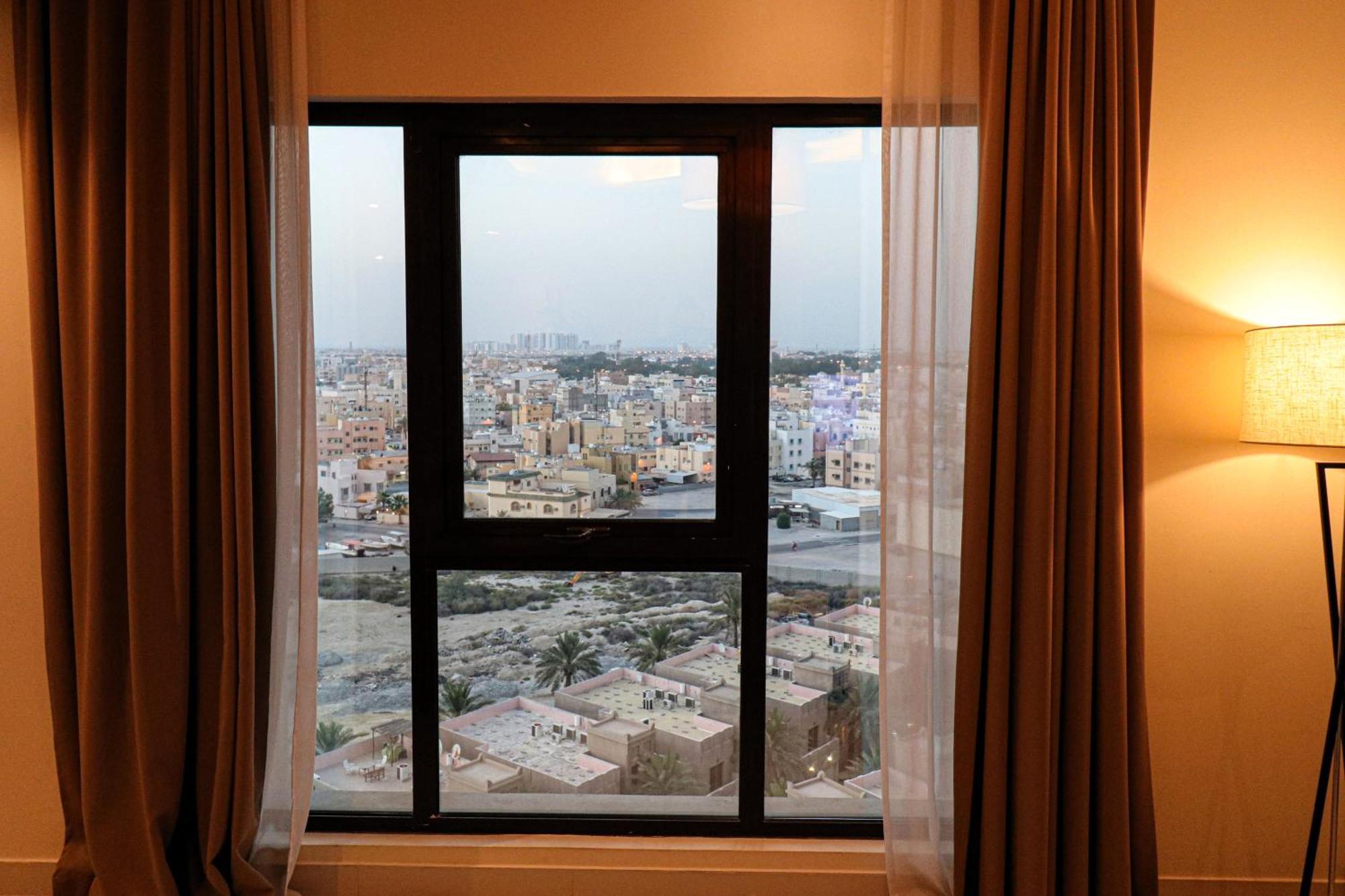 Lagoona Beach Luxury Resort And Spa Manama Luaran gambar The photo shows a window with a black frame and sheer curtains. Through the window, there is a view of a cityscape, displaying a densely populated area with various buildings and rooftops. The sky appears slightly overcast, suggesting a muted lightin