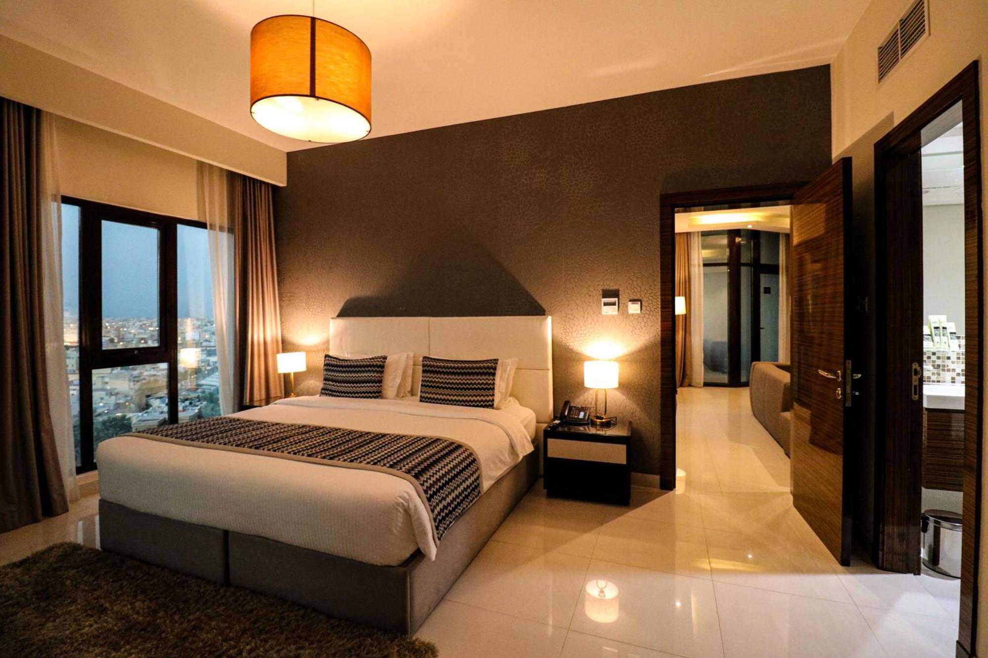 Lagoona Beach Luxury Resort And Spa Manama Luaran gambar The photo depicts a modern hotel room or suite. It features a large bed with a white and gray color scheme, accented with decorative pillows. On either side of the bed, there are sleek bedside lamps providing warm lighting. The walls are a dark color