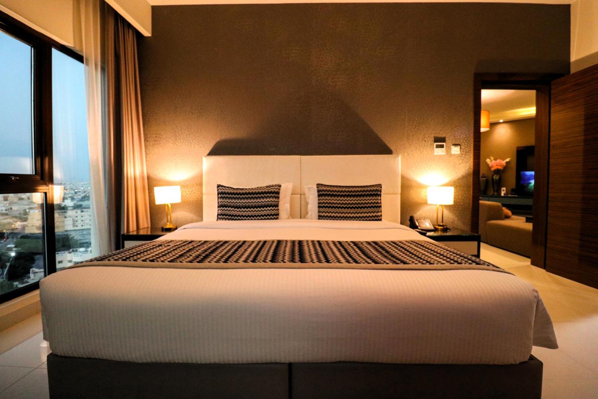 Lagoona Beach Luxury Resort And Spa Manama Luaran gambar The photo depicts a modern hotel room featuring a large bed with a white comforter and two decorative pillows. The headboard is light-colored, and the walls are painted a dark hue. On either side of the bed, there are bedside tables with lamps. The r