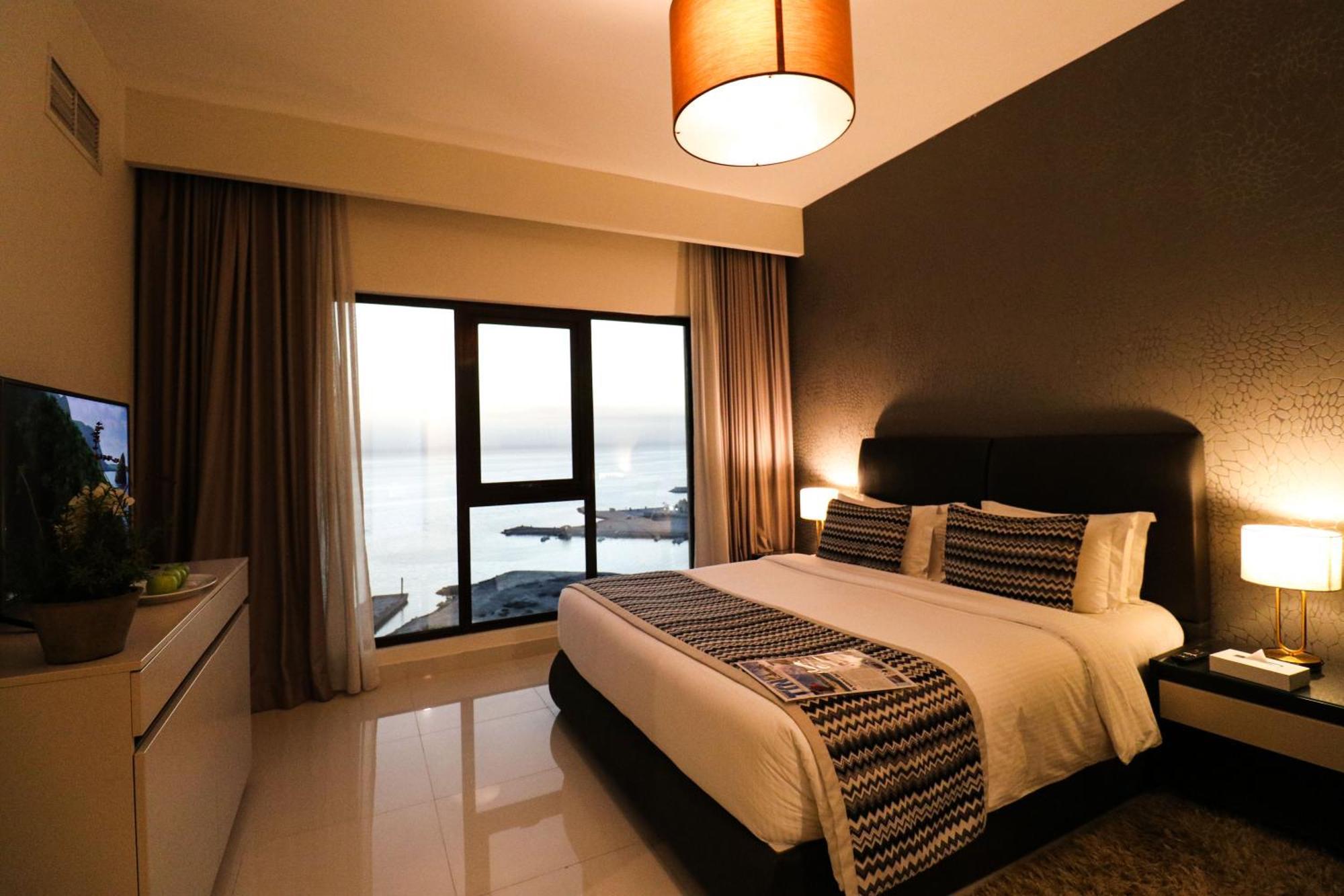 Lagoona Beach Luxury Resort And Spa Manama Luaran gambar The photo shows a modern hotel room featuring a neatly made bed with decorative pillows. The room has a stylish design with a dark accent wall and a large window that offers a view of the outdoors. Natural light comes in, giving the space a bright an
