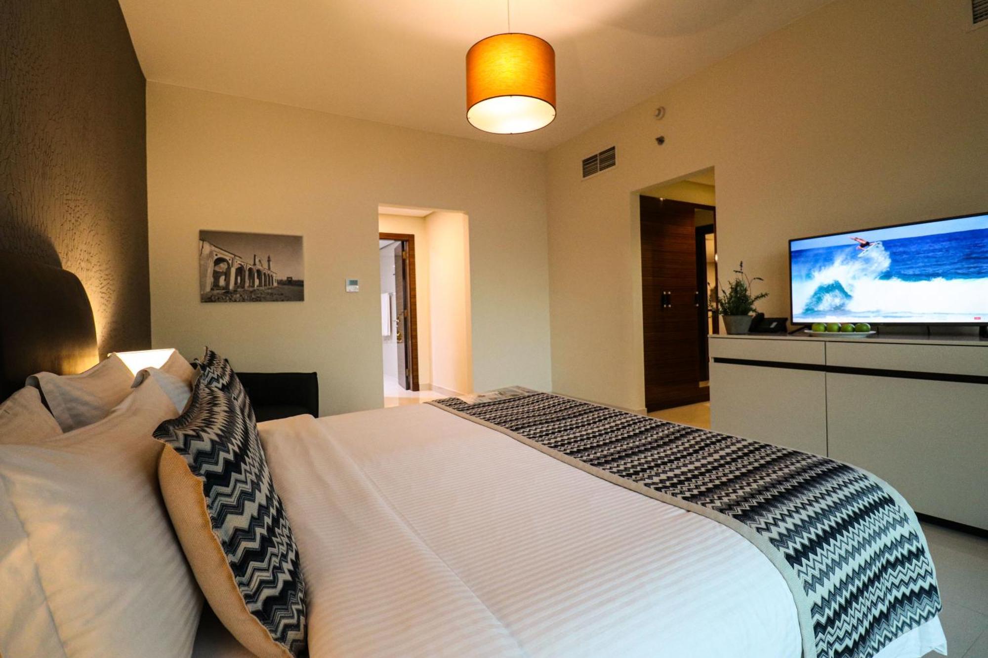 Lagoona Beach Luxury Resort And Spa Manama Luaran gambar The photo shows a well-decorated hotel room. There is a large bed with white linens and decorative pillows featuring a zigzag pattern. On the wall beside the bed is a piece of artwork, and there is a modern television on a white furniture unit opposi