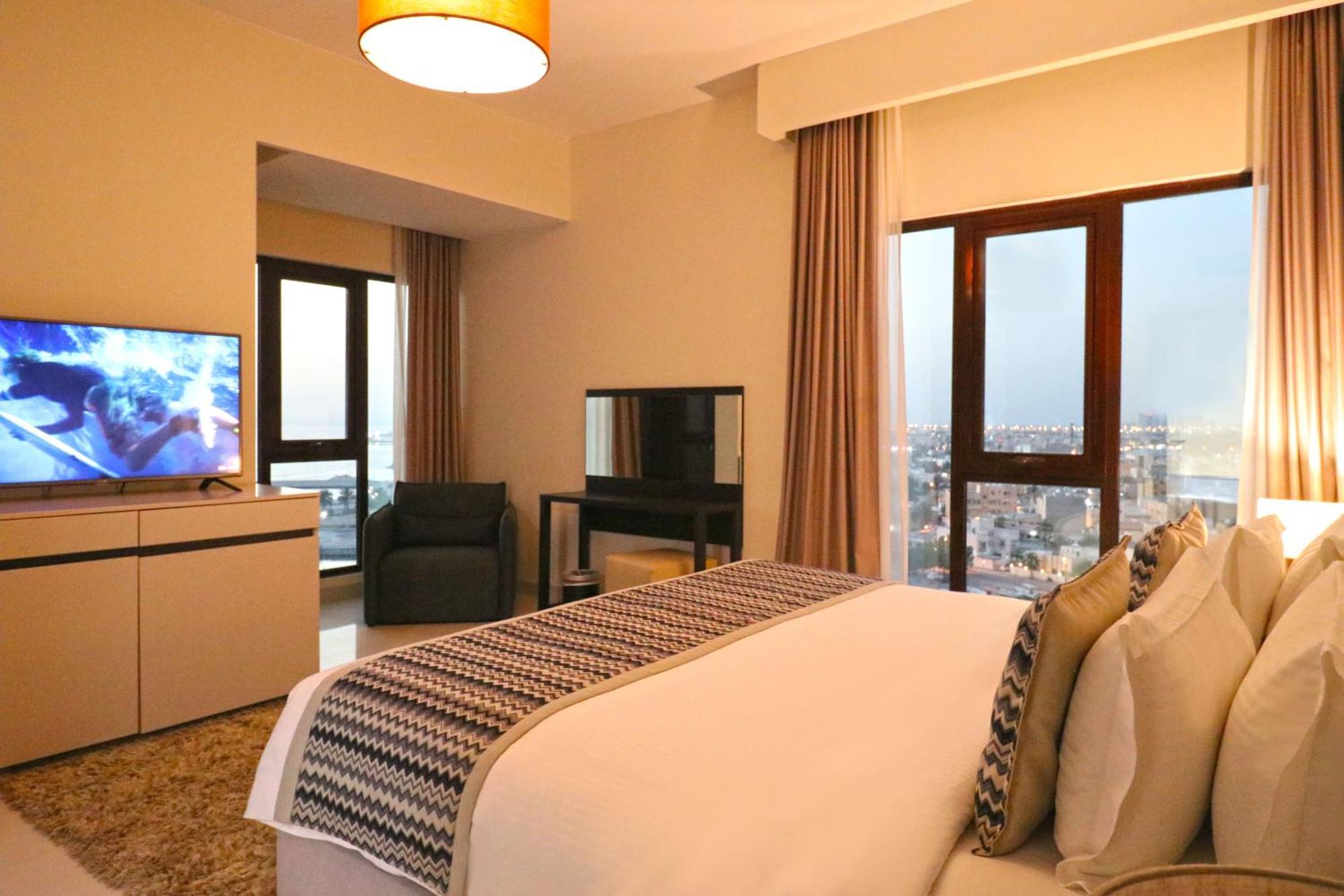 Lagoona Beach Luxury Resort And Spa Manama Luaran gambar The photo shows a modern hotel room or suite. It features a neatly made bed with decorative pillows in the foreground. To the left, there is a TV on a cabinet, and further back, a comfortable chair is situated in front of a large window that likely o