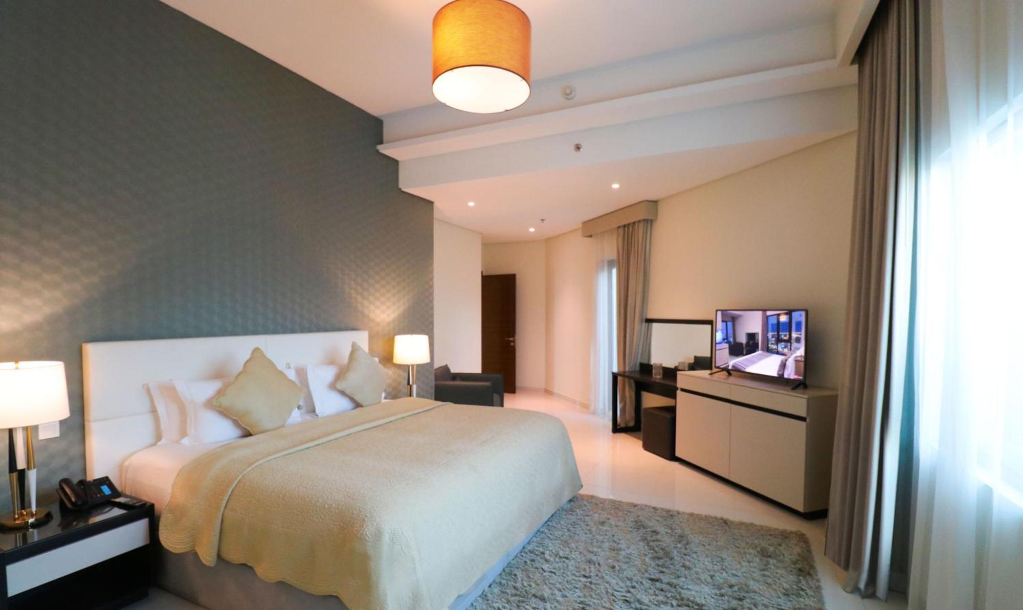 Lagoona Beach Luxury Resort And Spa Manama Luaran gambar The photo shows a modern hotel room featuring a large bed with a light-colored blanket and decorative pillows. The walls are painted in a subtle shade, and there is a textured accent wall behind the bed. On one side, a bedside lamp provides warm ligh